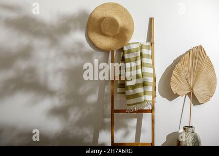 Strandtuch aus Bio-Leinen auf Holzleiter, Strohhut und getrocknetes Palmblatt an der weißen Wand mit Schatten von Baumblättern Stockfoto