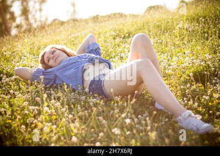 Junges blondes Landmädchen in Denim-Hemd mit Knöpfen, das heiße jean-Shorts trägt, posiert im Sommer zur goldenen Stunde im Freien und schaut verführerisch auf die Kamera Stockfoto