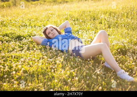 Junges blondes Landmädchen in Denim-Hemd mit Knöpfen, das heiße jean-Shorts trägt, posiert im Sommer zur goldenen Stunde im Freien und schaut verführerisch auf die Kamera Stockfoto