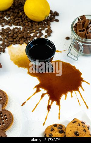 Verschütteter Kaffee aus einer schwarzen Tasse auf einem weißen Tisch. Kaffeebohnen, Zimt und Kekse. Frühstück in der Küche. Stockfoto