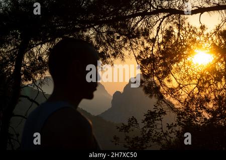 Silhouette des Menschen vor der malerischen Aussicht auf die Berge gegen den Himmel während des Sonnenuntergangs, durch Bäume. Stockfoto