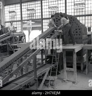 1950s, historisch, an der Vorderseite eines großen Lagerdepots oder Aufhängers männliche Arbeiter stehen auf einer Holzplattform und stapeln neu hergestellte Steine in der Ziegelei der London Brick Company, Stewartby, Bedford, England, Großbritannien. Mitte 1930s befand sich auf dem Gelände der größte Ofen der Welt, der 500 Millionen Ziegelsteine pro Jahr produzierte. Es gab 167 Schornsteine an den Werken. Stockfoto