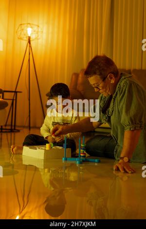 Enkelkind mit Großmutter, die am Abend vor dem Schlafengehen auf dem Boden sitzt und ein Brettspiel spielt. Glückliche Momente für die Familie. Stockfoto