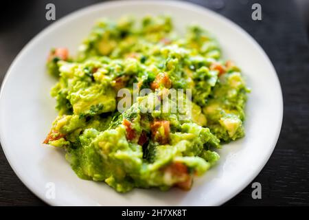 Oben Makro Nahaufnahme von Guacamole Gericht auf weißem Teller mit reifen grünen Avocado, Knoblauchtomaten und Koriander Stockfoto