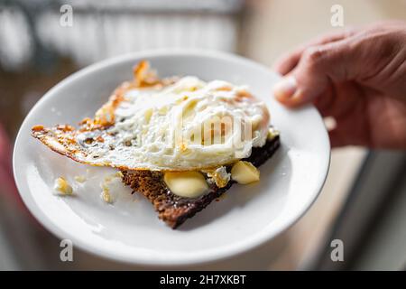 Makro-Nahaufnahme von Hand mit hausgemachtem, gebratenem, frischem Ei mit knusprigem Weiß auf geschmolzener Butter auf Toast-Roggenbrot Stockfoto