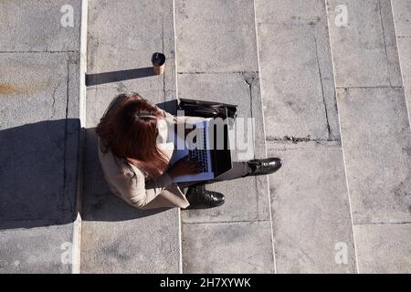 Draufsicht auf eine junge Frau, die mit einem Laptop auf einer Leiter sitzt. Stockfoto