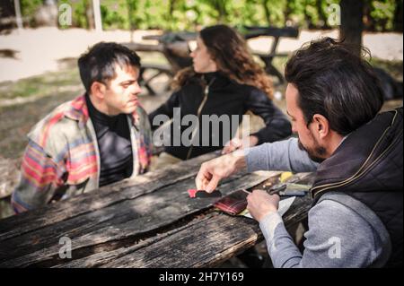 Gruppe von Freunden, die Hashish-Joint-Rolling-Marihuana-Zigarette für das Rauchen auf der Bank im öffentlichen Park im Freien vorbereiten. Junge Menschen rauchen Cannabis thc Gras Stockfoto