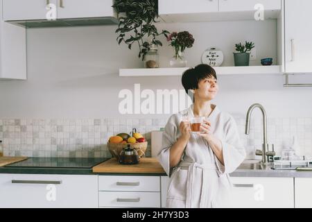 Fröhliche Frau im Bademantel hält eine Tasse frischen Tee in ihrer modernen weißen Küche. Konzept „Guten Morgen“. Positive Stimmung. Selektiver Fokus. Speicherplatz kopieren Stockfoto