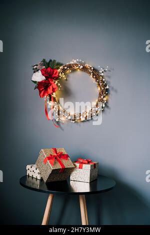 Moderner Weihnachtskranz aus gewebten Zweigen, Perlen und Lichtern an der grauen Wand über dem Tisch mit Geschenkschachteln. Minimalistisches festliches Heim dez Stockfoto