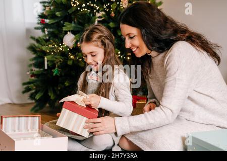 Aufgeregte Mutter und Tochter sitzen zu Hause in der Nähe von schön dekorierten Weihnachtsbaum und genießen das Öffnen ihrer Geschenke. Alleinstehende Elternzeit Stockfoto