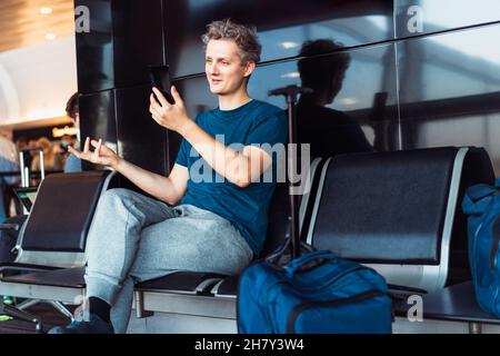 Junger männlicher Reisender wartet auf seinen Flug im Flughafen und hat ein Videotreffen. Anrufen, chatten mit seinem Smartphone. Digitaler Nomade, freiberuflicher Lebensstil. Stockfoto