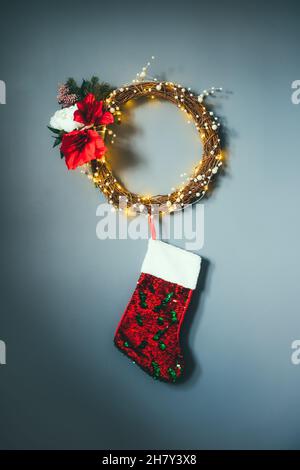 Moderner Blumenkranz aus gewebten Ästen, Perlen und Leuchten mit hängenden roten Pailletten-Weihnachtsstrümpfen an der grauen Wand. Minimalistische, festliche Einrichtung Stockfoto