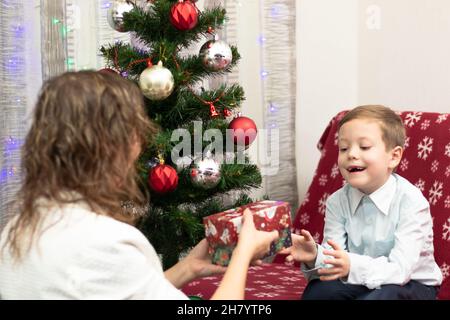 Mutter schenkt ihrem siebenjährigen Sohn zum Neujahr Geschenke in Schachteln vor dem Hintergrund eines Weihnachtsbaums zu Hause. Selektiver Fokus. Hochformat Stockfoto