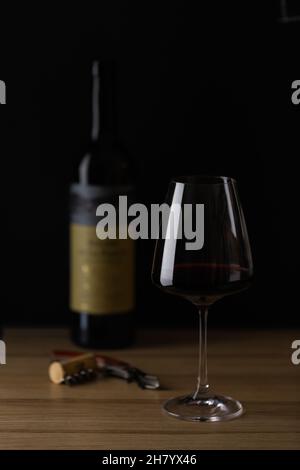 Der Wein wird in ein Glas gegossen und steht auf dem Tisch, neben der Weinflasche, Korkenzieher und Korken. Dunkler Hintergrund. Vorderansicht. Stockfoto