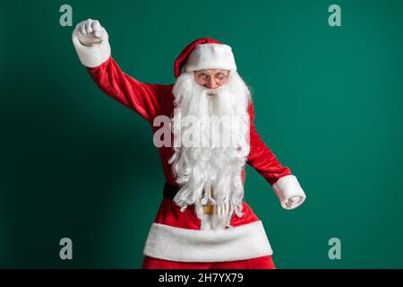 Lustiger Schauspieler im Kostüm des Weihnachtsmannes mit langem Bart gestikulieren beim Tanzen auf grünem Hintergrund Stockfoto