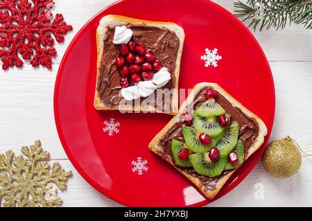 Kreative, festliche Sandwiches in Form eines Weihnachtsbaums und Weihnachtsmützen, die mit frischen Früchten und Salmellows auf einer roten Platte auf einem weißen Holzteller zubereitet werden Stockfoto