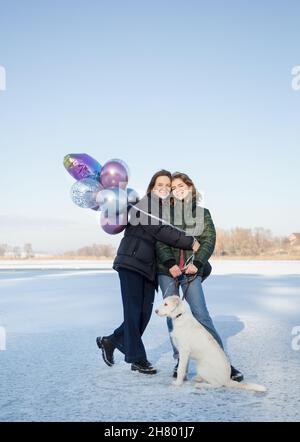 Zwei Frau mit weißem Hund und Haufen Ballons. Spaßspaziergang im Winter im Schnee. Lächelnde Erwachsene Tochter etwa 17-18 Jahre alt und Mutter etwa 40 Jahre alt Stockfoto