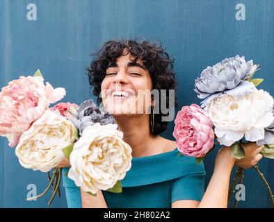 Entzückte junge Frau mit Blumensträußen duftender blühender Pfingstrosen, lachend mit breitem Lächeln und mit Blick auf die Kamera gegen die blaue Wand Stockfoto