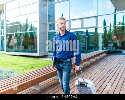 Ernsthafter bärtiger Arbeiter mit Netbook, der während einer Geschäftsreise neben einem Gebäude mit Glaswänden mit Koffer stand Stockfoto