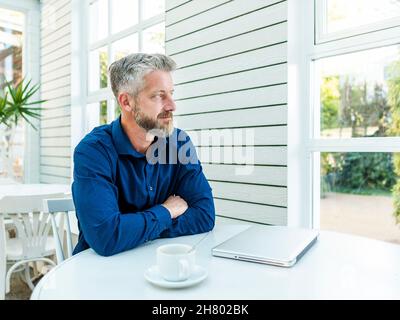 Seriöse bärtige männliche Fernarbeiterin faltete die Hände auf den Tisch mit Kaffee und Laptop und schaute nachdenklich weg Stockfoto