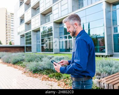 Fokussierter bärtiger Mann in Hemd und Jeans scrollendes Tablet, während er sich im Park in der Nähe eines modernen Bürogebäudes mit Arbeitsthemen fernab auseinandersetzt Stockfoto