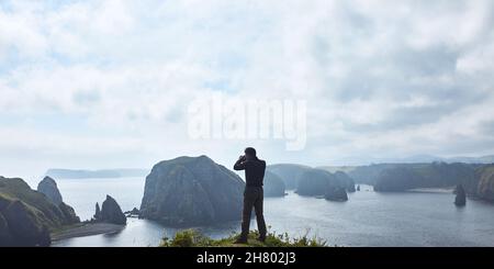 Ein männlicher Fotograf fotografiert vor dem Hintergrund einer wunderschönen Bucht. Die Hintergrundbeleuchtung und der große Raum an Himmel und Bucht schaffen eine ungewöhnliche Atmosphäre Stockfoto