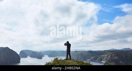 Ein männlicher Fotograf fotografiert vor dem Hintergrund einer wunderschönen Bucht. Die Hintergrundbeleuchtung und der große Raum an Himmel und Bucht schaffen eine ungewöhnliche Atmosphäre Stockfoto