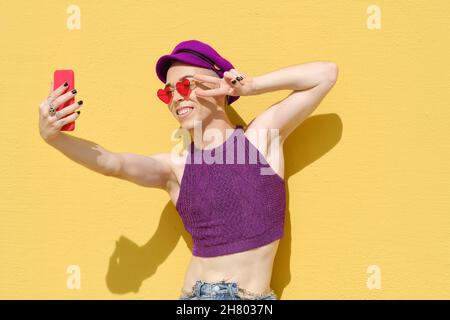 Nicht binäre junge Person, die Selfies mit einem Mobiltelefon macht, während sie an einer gelben Wand steht. Stockfoto