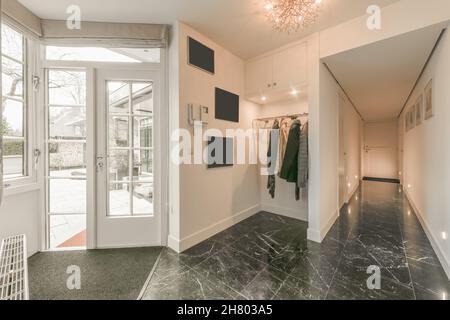 Innenansicht des Flurs mit weißen Wänden und gefliestem Boden und Kleiderbügel in der Nähe von Glastüren in einem modernen Apartment Stockfoto
