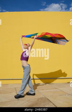 Glückliche junge nicht-binäre Person hält und winkt eine Regenbogenfahne im Freien. Stockfoto
