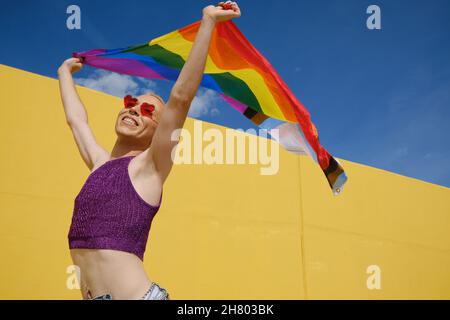 Junge nicht-binäre Person, die glücklich und aufgeregt aussieht, während sie draußen eine Regenbogenfahne schwenkt. Stockfoto