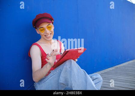 Lächelnder junger, nicht-binärer Mann in Baskenmütze und Sonnenbrille mit einem digitalen Tablet, während er im Freien sitzt. Stockfoto
