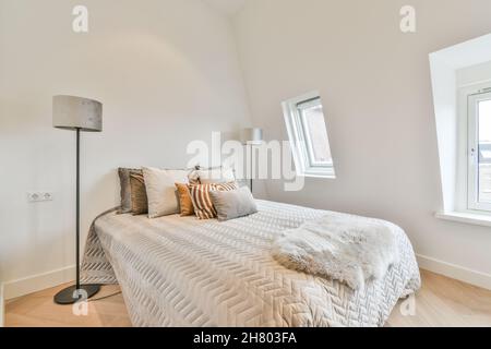 Komfortables Bett mit verschiedenen weichen Kissen, das an der weißen Wand zwischen hohen Stehlampen im hellen, modernen Schlafzimmer des Apartments platziert ist Stockfoto