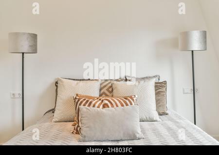 Komfortables Bett mit verschiedenen weichen Kissen, das an der weißen Wand zwischen hohen Stehlampen im hellen, modernen Schlafzimmer des Apartments platziert ist Stockfoto