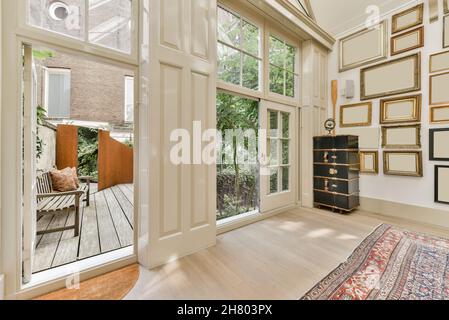 Weiße Wand mit verschiedenen leeren Bilderrahmen in einem hellen, geräumigen Zimmer mit Teppich in der Nähe von geöffneten Türen mit Blick auf die Terrasse mit grünen Bäumen Stockfoto