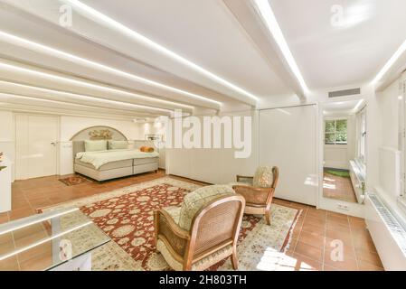 Komfortables Bett auf Teppich im hellen, geräumigen Schlafzimmer mit Fernseher in der Nähe von dekorativen Elementen und glühenden Lampen im Apartment Stockfoto