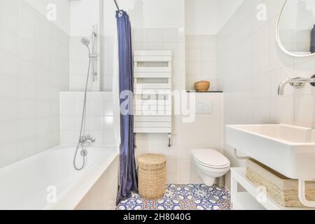 Weiße Badewanne mit Schlauch und Vorhang in der Nähe der Toilette im hellen modernen Badezimmer mit Waschbecken an der gefliesten Wand mit Spiegel platziert Stockfoto