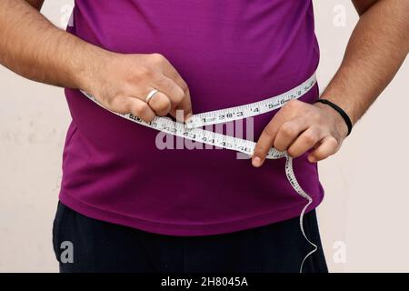 Ein asiatischer Mann misst seinen dicken Bauch mit einem Maßband auf einem schlichten Hintergrund Stockfoto