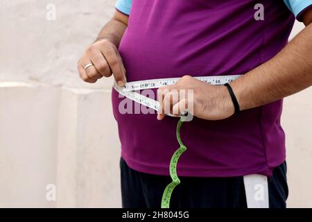 Ein asiatischer Mann misst seinen dicken Bauch mit einem Maßband auf einem schlichten Hintergrund Stockfoto