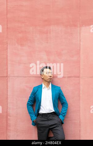 Selbstbewusster asiatischer Mann in formeller Kleidung in stilvoller blauer Anzugjacke, der wegschaut, während er sich an einer hellen Wand auf der Straße in der Stadt lehnt Stockfoto