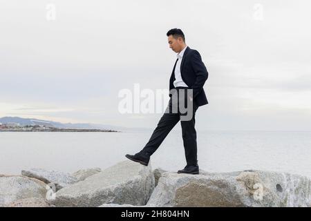 Ganzkörper-Seitenansicht des asiatischen männlichen Unternehmers in formeller Kleidung, der auf steinerner Küste nahe dem Meer in der Küstenregion läuft Stockfoto