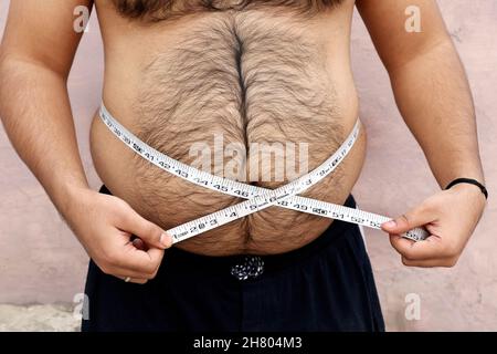 Ein asiatischer Mann misst seinen dicken Bauch mit einem Maßband auf einem schlichten Hintergrund Stockfoto