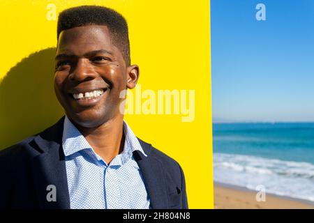 Positiver afroamerikanischer Unternehmer in formellem Anzug, der an der gelben Wand am Sandstrand mit wehendem Meer steht Stockfoto