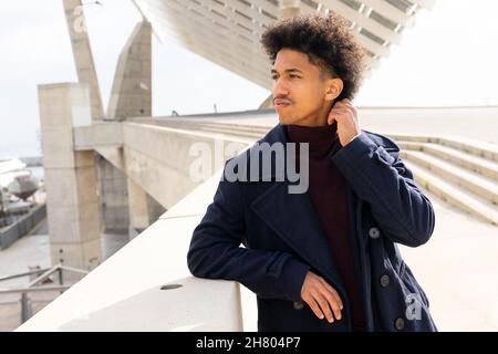 Nachdenklicher afroamerikanischer junger Mann in elegantem Mantel, der sich am Zaun lehnt und nachdenklich davonschaut Stockfoto