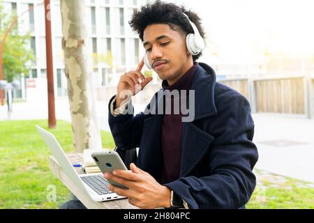 Konzentrierter afroamerikanischer Student im Mantel, der Musik in kabellosen Kopfhörern hört, während er im Park auf dem Handy surft und auf dem Laptop studiert Stockfoto