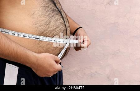 Ein asiatischer Mann misst seinen dicken Bauch mit einem Maßband auf einem schlichten Hintergrund Stockfoto