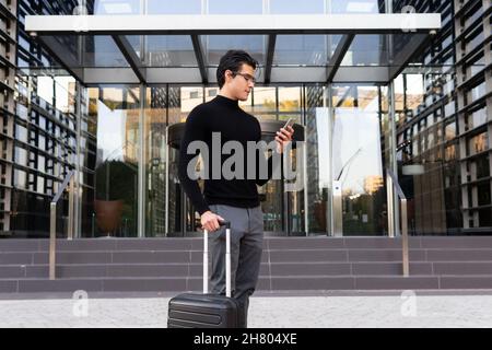 Seitenansicht des intelligenten asiatischen männlichen Unternehmers in stilvollem Trage-Surfcellphone, während er mit Gepäck in der Nähe eines modernen Gebäudes in der Stadt steht Stockfoto