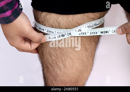 Ein erwachsener Mann misst seinen Oberschenkel mit einem Maßband auf einem schlichten Hintergrund Stockfoto