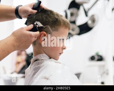 Anonymous Crop professionellen Friseur in Schürze schneiden Haare von Jungen in Cape in professionellen Salon eingewickelt Stockfoto