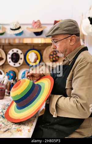 Ältere männliche Meister in Schürze Malerei Regenbogenmuster auf stilvollen Strohhut mit Pinsel stehen an der Werkbank mit bunten Farbstoffen Stockfoto